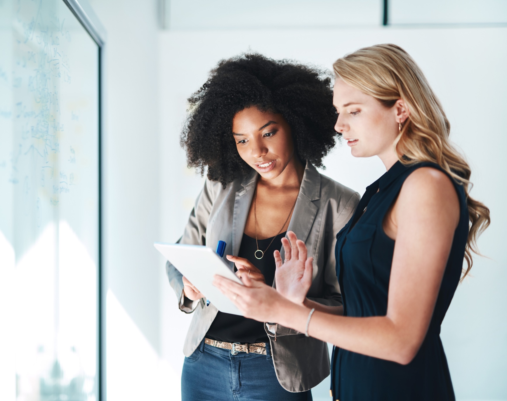 Two women reviewing a tablet and writing on a whiteboard